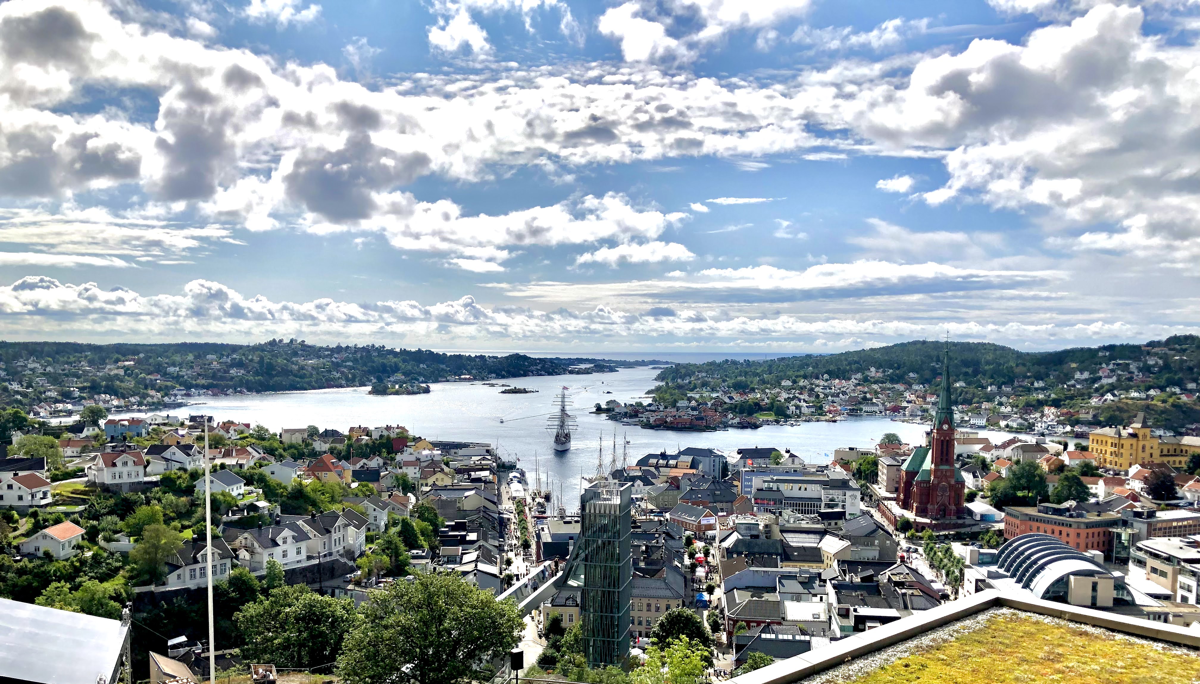 Panoramautsikt over Arendal sentrum og Galtesund med småbåthamn, historiske bygningar og kyrkje i forgrunnen, omgitt av sjø, øyar og kupert landskap under delvis skydekt himmel.
